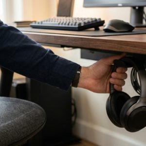 Hand reaching under a clean office desk to grab headphones hanging from a sturdy mount.